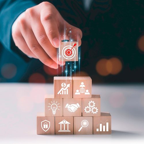 A imagem mostra uma mão masculina montando uma pilha de elementos fundamentais para a estratégia adaptativa. Na mão se encontra a peça com o símbolo de um alvo para representar o foco.