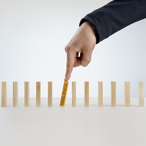 A imagem mostra uma mão masculina, especificamente o dedo indicador, segurando uma peça de madeira que está no meio de uma fileira de peças, evitando que ela caia. Na peça tem escrito o nome resiliência.