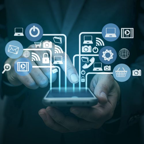 Mãos masculinas seguram um celular com a tela virada para cima. Da tela saem diversos ícones como telefone, computador, símbolo de conexão por wi-fi, carta, engrenagem, computador, entre outros. A imagem possui vários tons de azul e também branco.