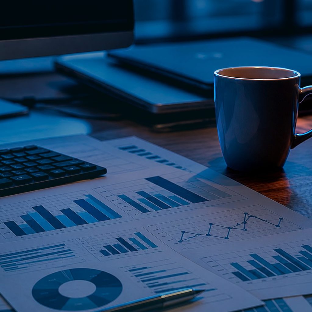 Mesa de trabalho com gráficos financeiros impressos, teclado, notebook e uma caneca de café ao lado, representando análise de dados e gestão empresarial.
