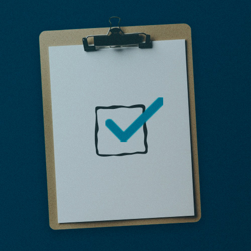 A imagem mostra uma prancheta (clipboard) com uma folha de papel branca presa por um clipe metálico na parte superior. No centro da folha há um ícone de caixa de seleção com um grande símbolo de check azul, representando aprovação, conformidade, verificação ou conclusão de uma tarefa. O fundo é azul-escuro, criando contraste com os elementos claros da imagem.